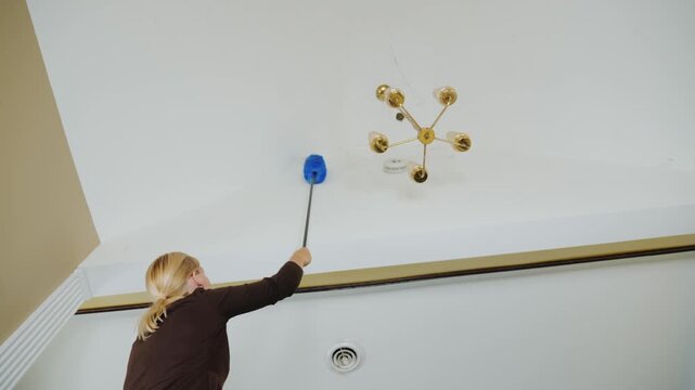 Woman dusting high ceiling and chandelier with long duster pole on ladder.