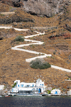 A zigzag white path climbs the rocky hillside toward a traditional windmill on Thirassia Island near Santorini, Greece, a popular swimming stop on catamaran tours.