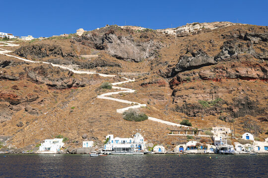 Whitewashed buildings and a winding trail rise from the sea along the rugged coast of Thirassia Island near Santorini, Greece, under clear Mediterranean skies.