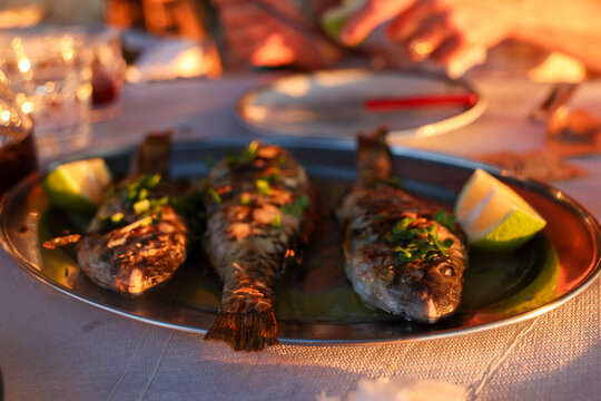 Three grilled fish garnished with herbs and lime wedges are served on a platter in Greece, glowing in warm evening light.