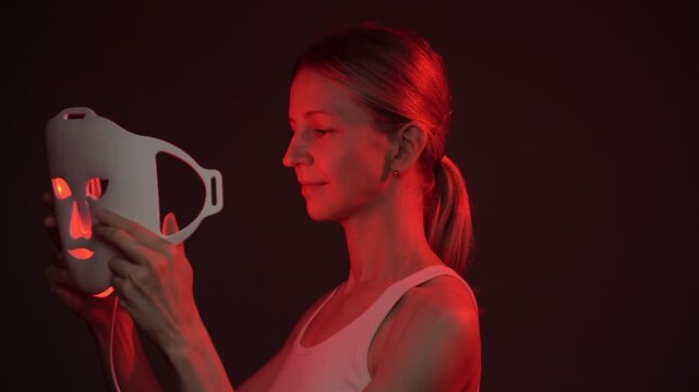woman using an infrared facial mask. profile side view then turns smiling and looking at camera.  Professional cosmetic skin care. vertical video footage black background studio shoot, contrast red li