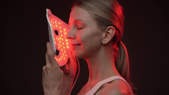 beautiful woman looks at the camera and smiles, then turns in profile to an infrared cosmetic mask. biohacker theme professional skin care video footage. black background