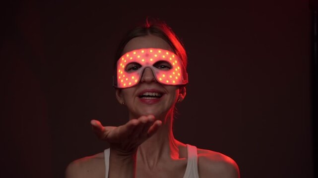 Red and infrared light therapy eye mask. smiling woman wearing infrared light therapy eye mask. looking at the camera, smiling sending air kiss. dark background. video footage
