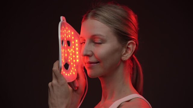 blonde woman holding infrared light therapy LED mask and looking to the camera, after turning her face to the mask. black  background. Contrast red light. horizontal composition video footage