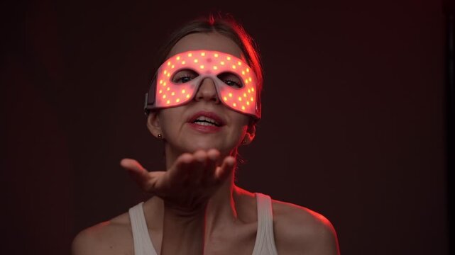 Red light therapy eye mask. smiling woman wearing infrared light therapy eye mask. looking at the camera, smiling sending air kiss. dark background. video footage