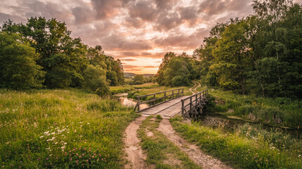 Sendero de tierra que atraviesa un puente de madera entre árboles y praderas, iluminado por la luz del atardecer bajo un cielo con nubes, evocando paz y naturaleza. © Argiduna