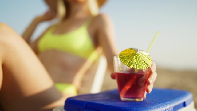 Refreshing pink cocktail with drinking straw and colorful paper umbrella on beachside table, with blurred woman relax on sun lounger in background. Summer vacation,tropical refreshment,seaside leisure