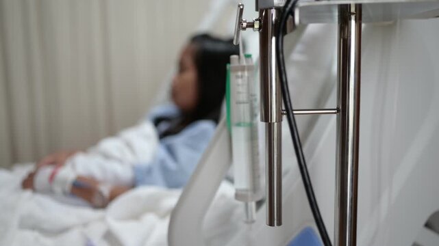 Female patient sits on a hospital bed beside an IV infusion pump while receiving medical care. Concept of hospitalization, healthcare, patient recovery, treatment, and clinical care.