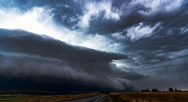 Dramatic landscape featuring a massive shelf cloud of a supercell thunderstorm moving over a rural road and countryside fields, capturing the powerful atmosphere of an approaching storm.