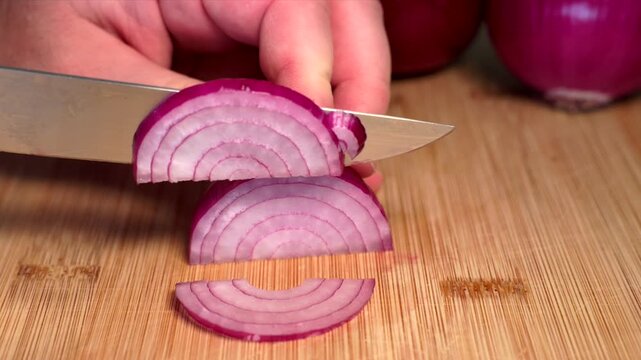 A hand is holding a red onion on a cutting board and slicing it with a knife. Contains vitamin C and fiber. Nutrition, vegetables, healthy lifestyle, fresh, raw food. 
