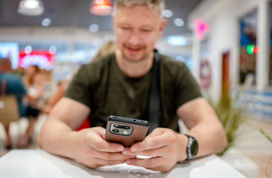 Middle aged man sits at table checking his smartphone screen inside a bright shopping center
