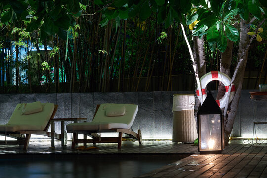 Relaxing poolside lounge area at night featuring comfortable sunbeds, a decorative lantern, and a lifebuoy against a bamboo fence.