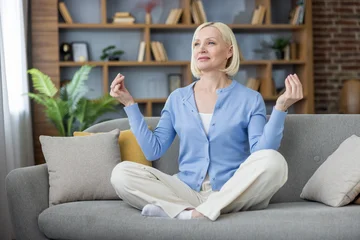 Fotobehang Lotusbloem Mature woman sitting in lotus position on a sofa, practicing meditation and relaxation in a comfortable living room, finding mental clarity and spiritual harmony during wellness routine  © Liubomir