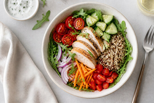 A vibrant, healthy bowl of sliced chicken, fresh vegetables, and quinoa is presented on a light surface.