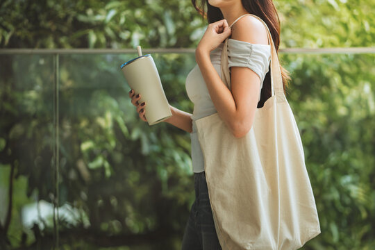 Side view close up of Asian woman holding reusable tumbler with straw and carrying cloth tote bag while walking outdoors near green park for eco friendly, zero waste and sustainable lifestyle concept.