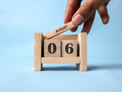 A woman's hand holds a wooden block with August written on it on a wooden block calendar for August 6th against a light blue background.