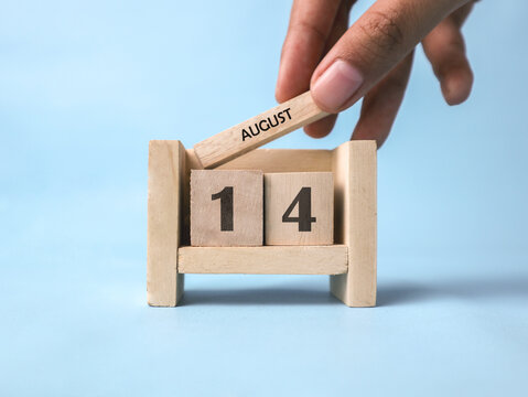 A woman's hand holds a wooden block with August written on it on a wooden block calendar for August 14th against a light blue background.