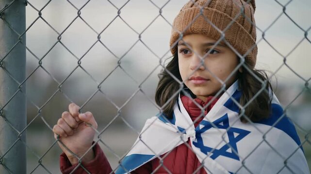 Young girl in brown beanie wearing Israeli flag standing behind chain link fence, close up shot