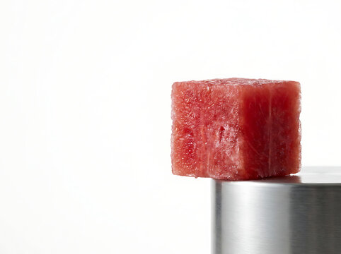 Creative close-up shot of a single vibrant red watermelon cube meticulously balanced on a sleek metallic surface against a clean white background