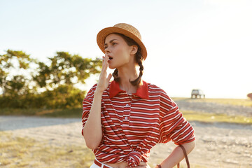 Obraz premium Young woman in a straw hat and striped shirt yawns while standing outdoors. Authentic lifestyle portrait of a girl posing with braided hair on a sunny summer day.