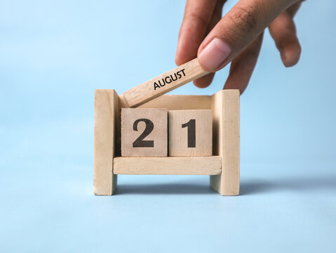 A woman's hand holds a wooden block with August written on it on a wooden block calendar for August 21st against a light blue background.