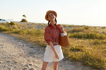 Obraz premium Happy woman enjoying lifestyle during a summer walk on a sandy beach. She wears a straw hat and striped shirt, holding a bag. Sunny outdoor scene with warm lighting.