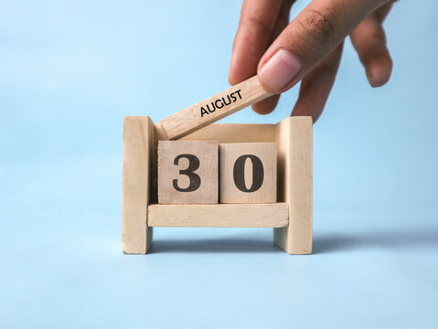 A woman's hand holds a wooden block with August written on it on a wooden block calendar for August 30th against a light blue background.