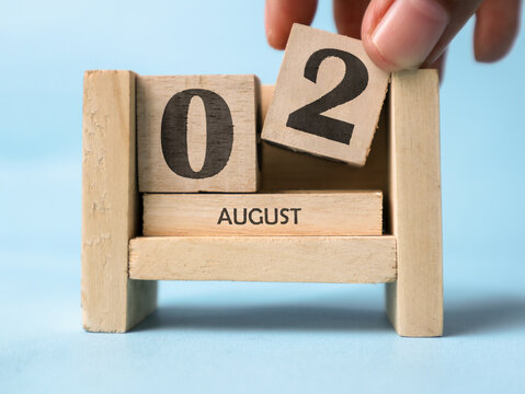 A woman's hand is holding a wooden block with the number 2 written on it on a wooden block calendar for August 2nd on a light blue background.