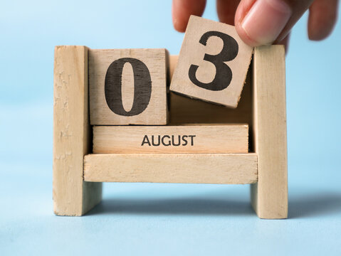 A woman's hand is holding a wooden block with the number 3 written on it on a wooden block calendar for August 3rd on a light blue background.