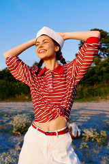 Obraz premium Smiling woman lifestyle posing outdoors in a striped shirt and hat. Sunny summer background, golden hour lighting. Concept of travel, adventure, freedom, and vacation mood.