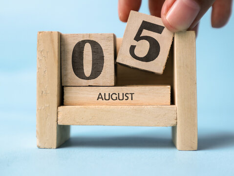 A woman's hand is holding a wooden block with the number 5 written on it on a wooden block calendar for August 5th on a light blue background.