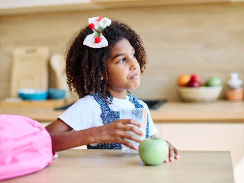 Young girl enjoying a glass of milk while sitting at a kitchen table with an apple nearby  preparing for school modern kitchen