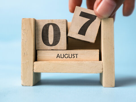 A woman's hand is holding a wooden block with the number 7 written on it on a wooden block calendar for August 7th on a light blue background.