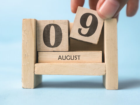 A woman's hand is holding a wooden block with the number 9 written on it on a wooden block calendar for August 9th on a light blue background.