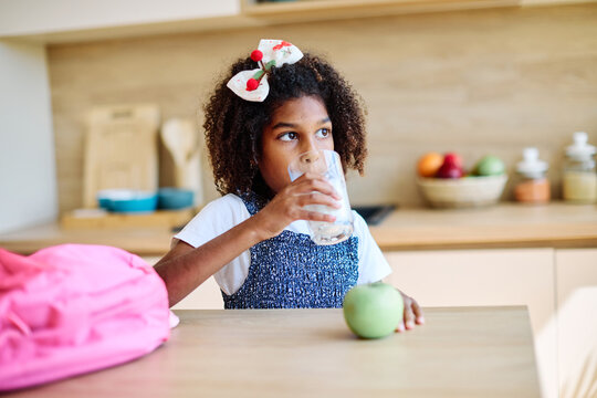 Young girl enjoying a glass of milk while sitting at a kitchen table with an apple nearby  preparing for school modern kitchen