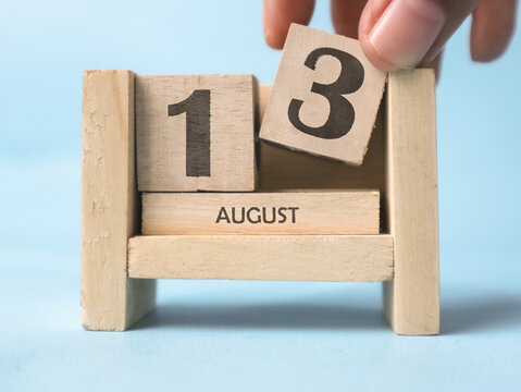 A woman's hand is holding a wooden block with the number 3 written on it on a wooden block calendar for August 13th on a light blue background.