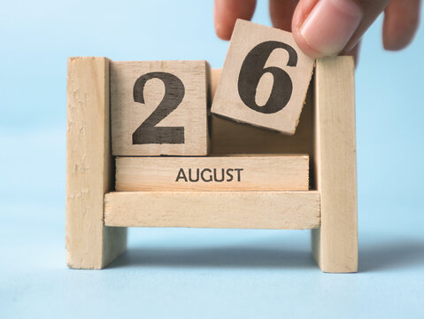 A woman's hand is holding a wooden block with the number 6 written on it on a wooden block calendar for August 26th on a light blue background.