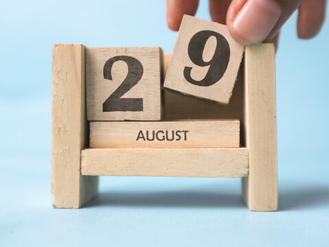 A woman's hand is holding a wooden block with the number 9 written on it on a wooden block calendar for August 29th on a light blue background.