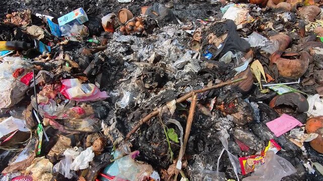 Video of Ash and charred waste lies scattered. Broken plastic and burnt packaging mix with coconut husks. Burned items rest near unburned coconuts and green grass. 4K Footage at a rubbish dump site
