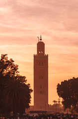 Obraz premium Tall minaret tower rises above crowded market below in the ornage light of sunset, Marrakesh Morocco