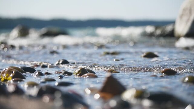 Shallow sea water gently flowing over smooth pebbles under bright daylight by rocky shore