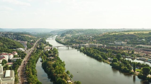 Vltava river flows through green urban landscape in Prague. Scenic riverside displays road traffic and distant bridges. Natural city environment serves as European tourism destination.