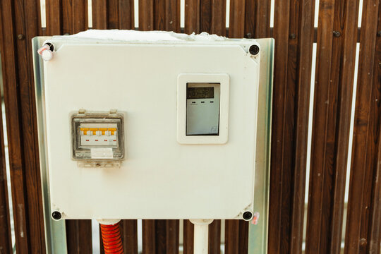 White electrical panel with meter and breakers on wooden fence, topped with snow, in bright outdoor light