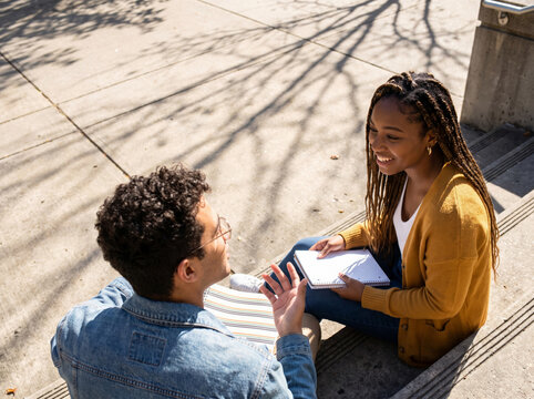 Two university students having an animated conversation on sunny concrete steps, representing authentic campus lifestyle and communication.