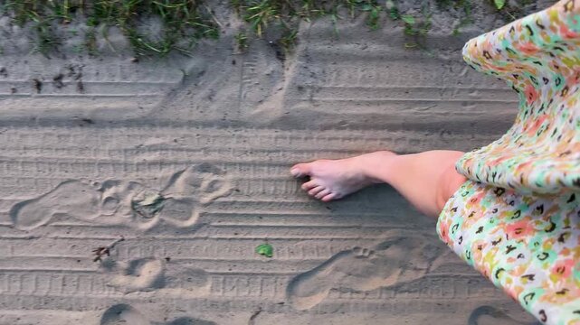A top-down perspective shows a woman in a floral dress walking barefoot along a sandy dirt road marked with footprints.
