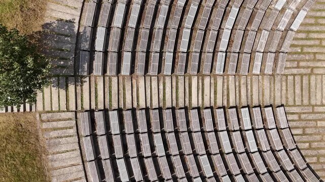 Top down aerial view slowly rotating above empty curved wooden benches and stone steps of an outdoor amphitheater in a public park, revealing geometric patterns