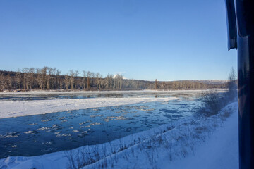View past the edge of a train carriage across an icy Alaskan river with drifting floes, frosted cottonwood trees and distant snowy mountains under a clear blue sky. © Benedikt