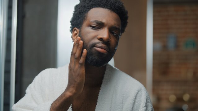 Portrait confident young African American man touching gentle face skin beard grooming looking at mirror reflection wearing bathrobe at bathroom handsome ethnic guy bearded male touch facial hairstyle