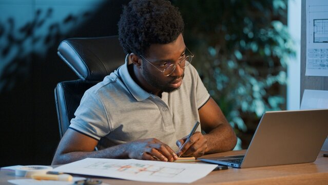 Concentrated African American business man architect analyzing construction blueprint writing notes with laptop at night office focused male guy businessman analyze architecture plan write at evening