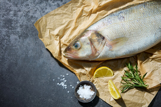 Raw fish, sea bass on parchment paper. Ingredients for cooking, grill, roasting. Top view. Grey background. Close up. Copy space.